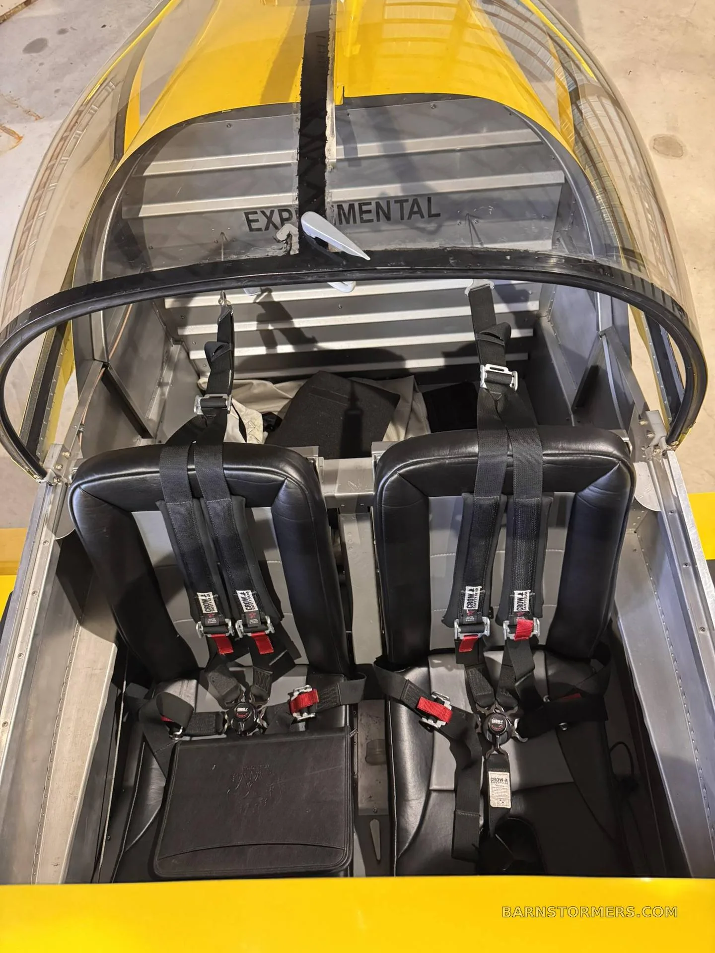 An overhead view into the cabin of a yellow experimental aircraft shows two black seats with harnesses and the word "EXPERIMENTAL" on the bulkhead.