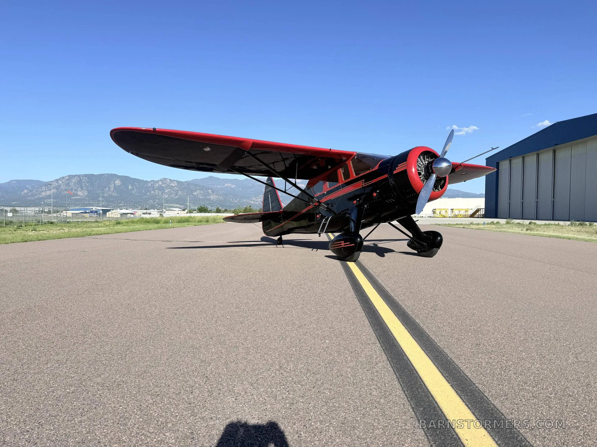 A black and red vintage high-wing monoplane with a radial engine, parked on an asphalt runway with mountains in the background.