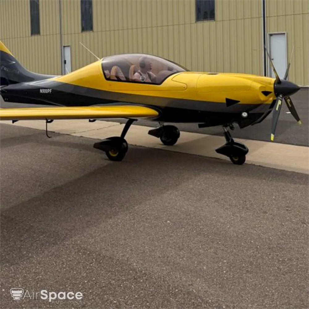 A yellow and black two-seat experimental aircraft with a bubble canopy is parked on an asphalt apron next to a yellow hangar.