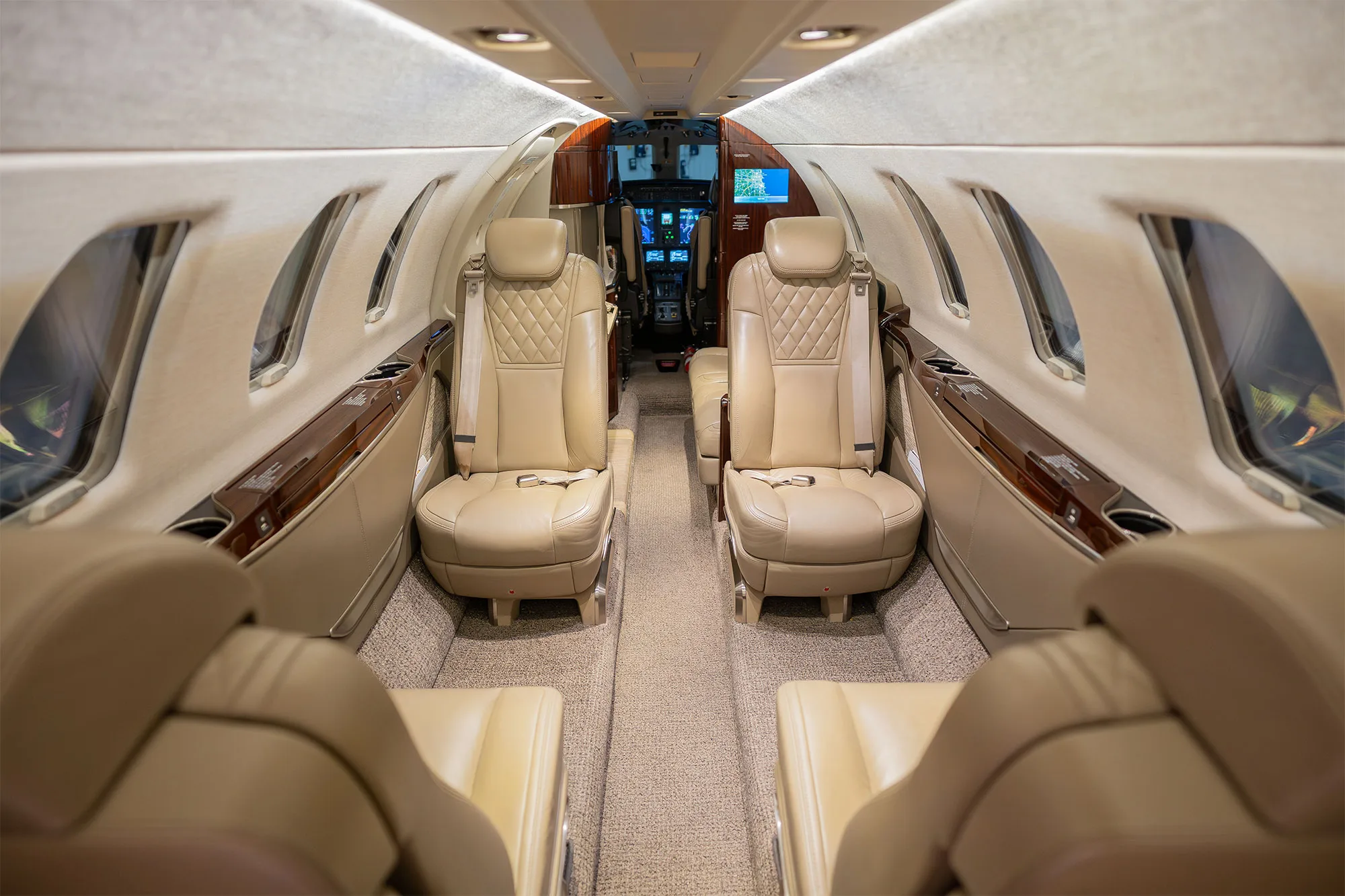 The luxurious interior cabin of a business jet, featuring beige leather seats and wood accents, looking towards the cockpit.