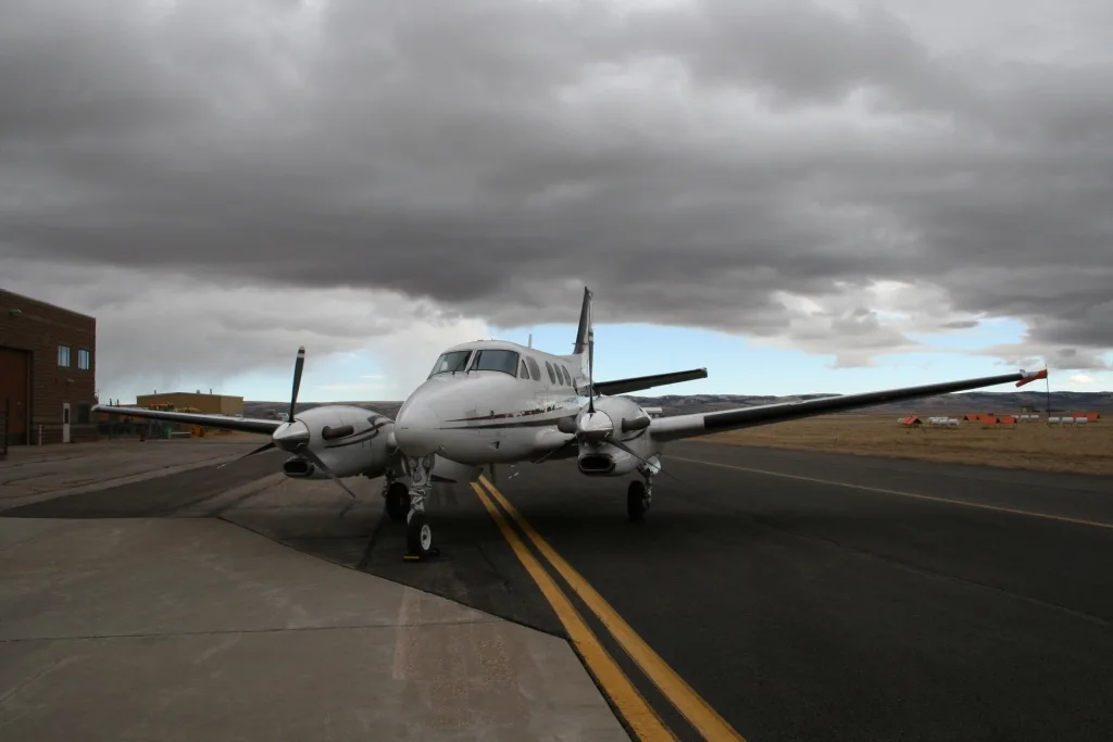 A white twin-engine turboprop aircraft with dark red and blue stripes, parked on an asphalt runway next to a building under a cloudy sky.