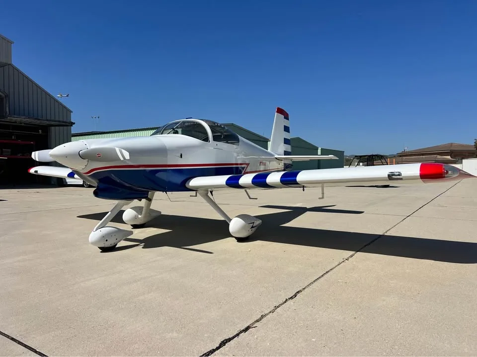 A white, blue, and red single-engine aircraft with a bubble canopy parked on an asphalt tarmac next to a hangar.