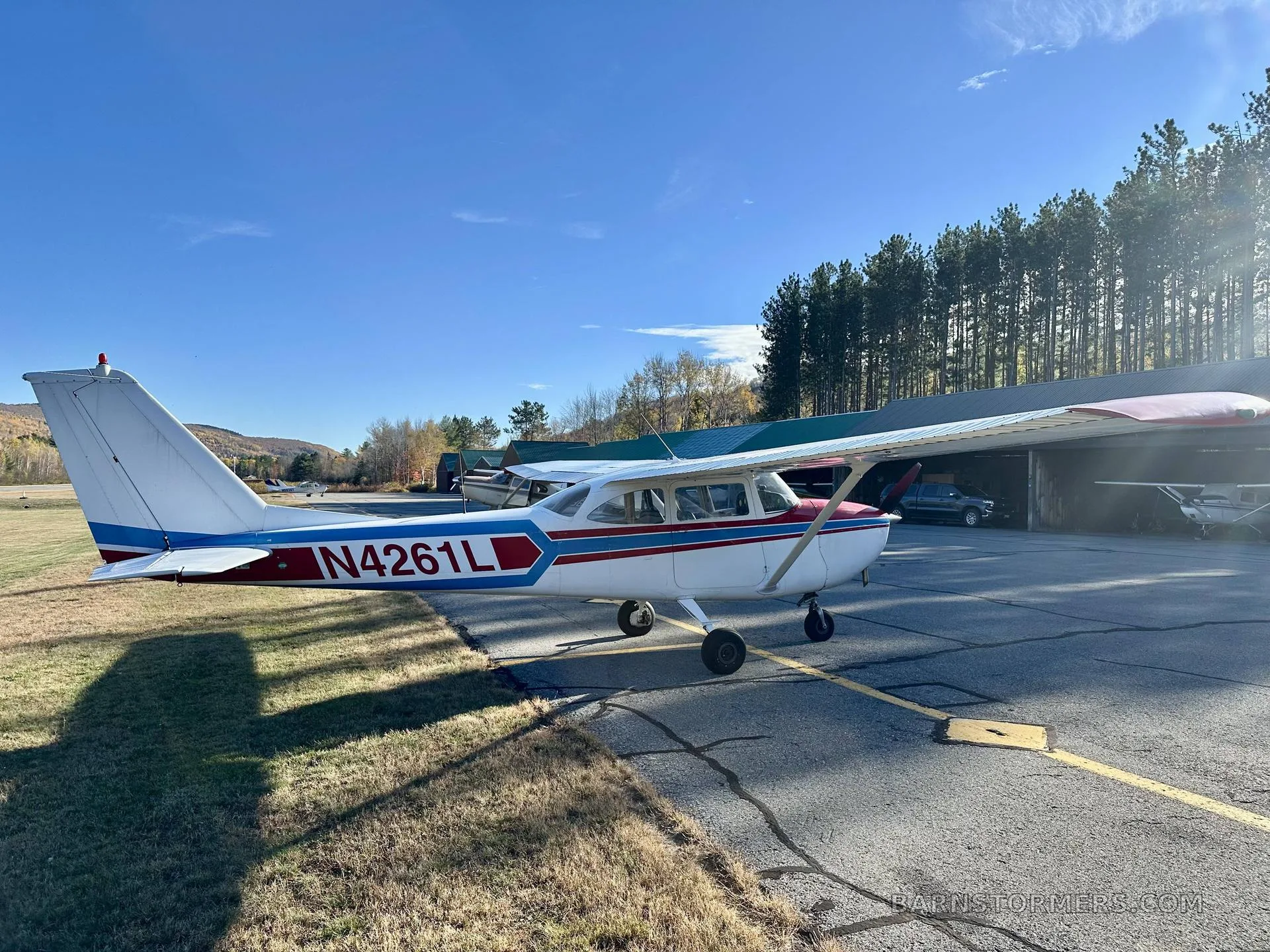 A white single-engine propeller aircraft with red and blue stripes, viewed from the rear-right, parked on an asphalt ramp next to a line of pine trees.