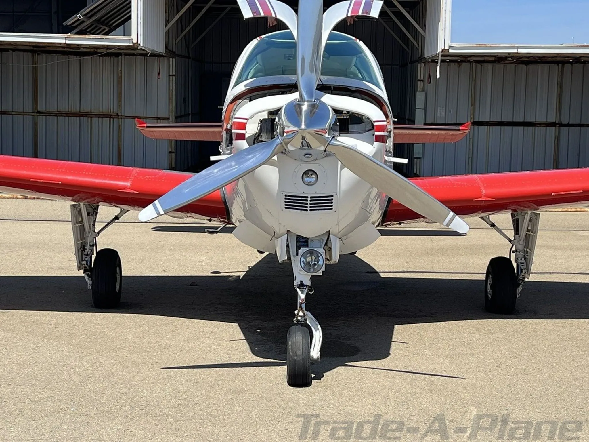 Frontal view of a white and red Beechcraft Bonanza with a polished propeller, parked in front of a hangar.