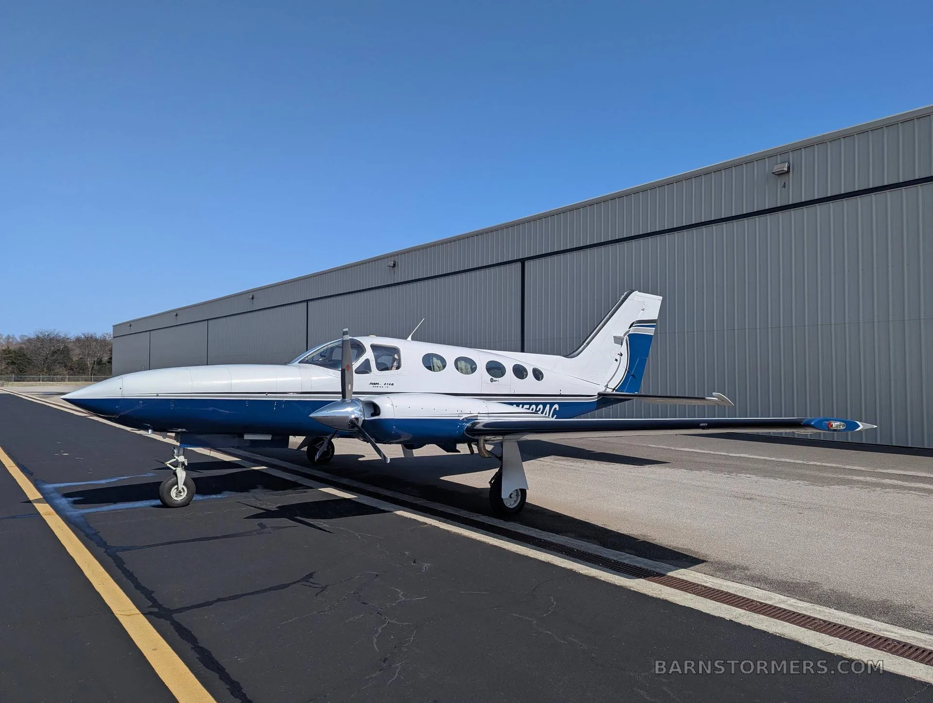 A white and blue twin-engine propeller aircraft (Cessna 414 Chancellor) with tail number N533AC parked on an asphalt ramp with yellow markings, viewed from the front-left side.