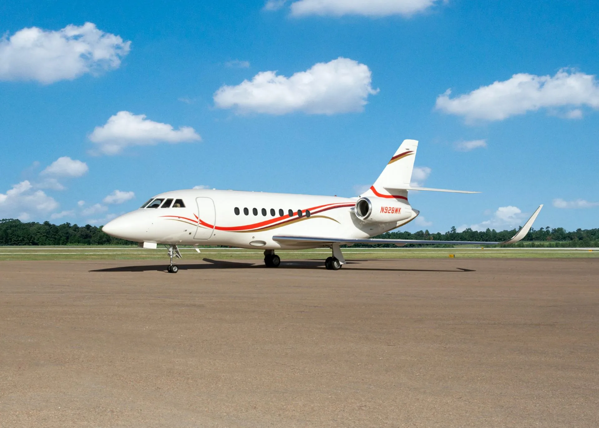 A white Dassault Falcon business jet with red and gold stripes is parked on an airfield under a blue sky.