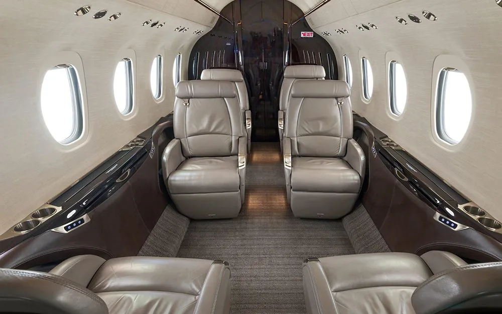 The interior cabin of a private jet, showing two rows of light brown leather seats facing each other.