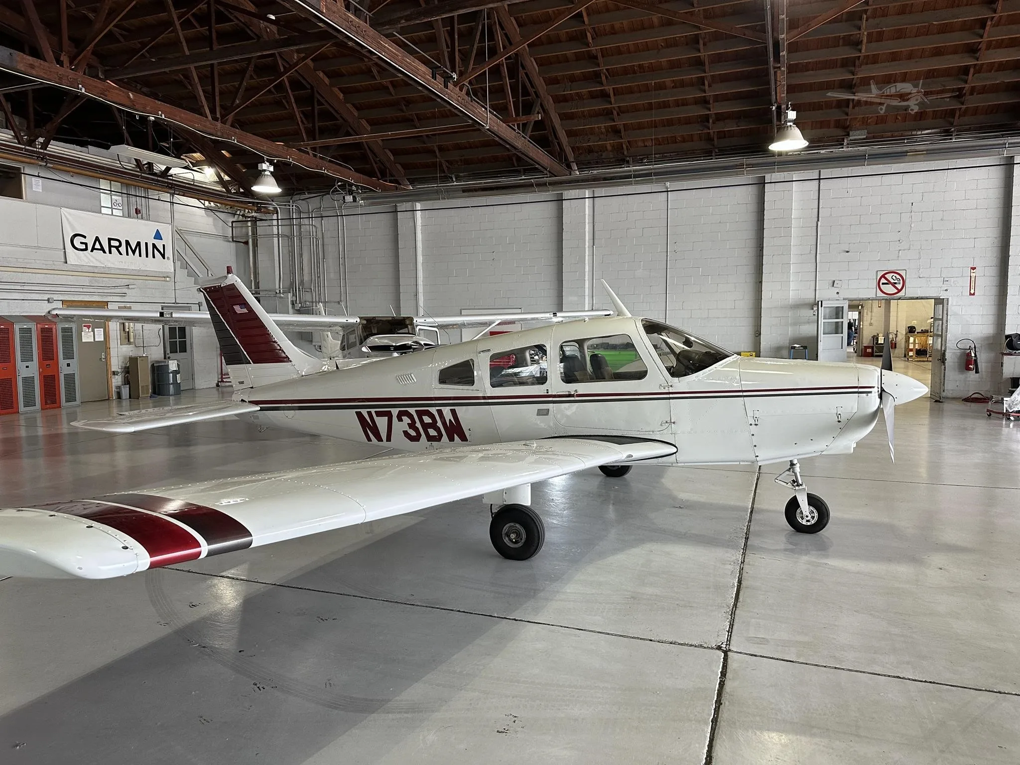 A white single-engine aircraft with maroon and black stripes, registration N73BW, viewed from the front-right in a hangar.