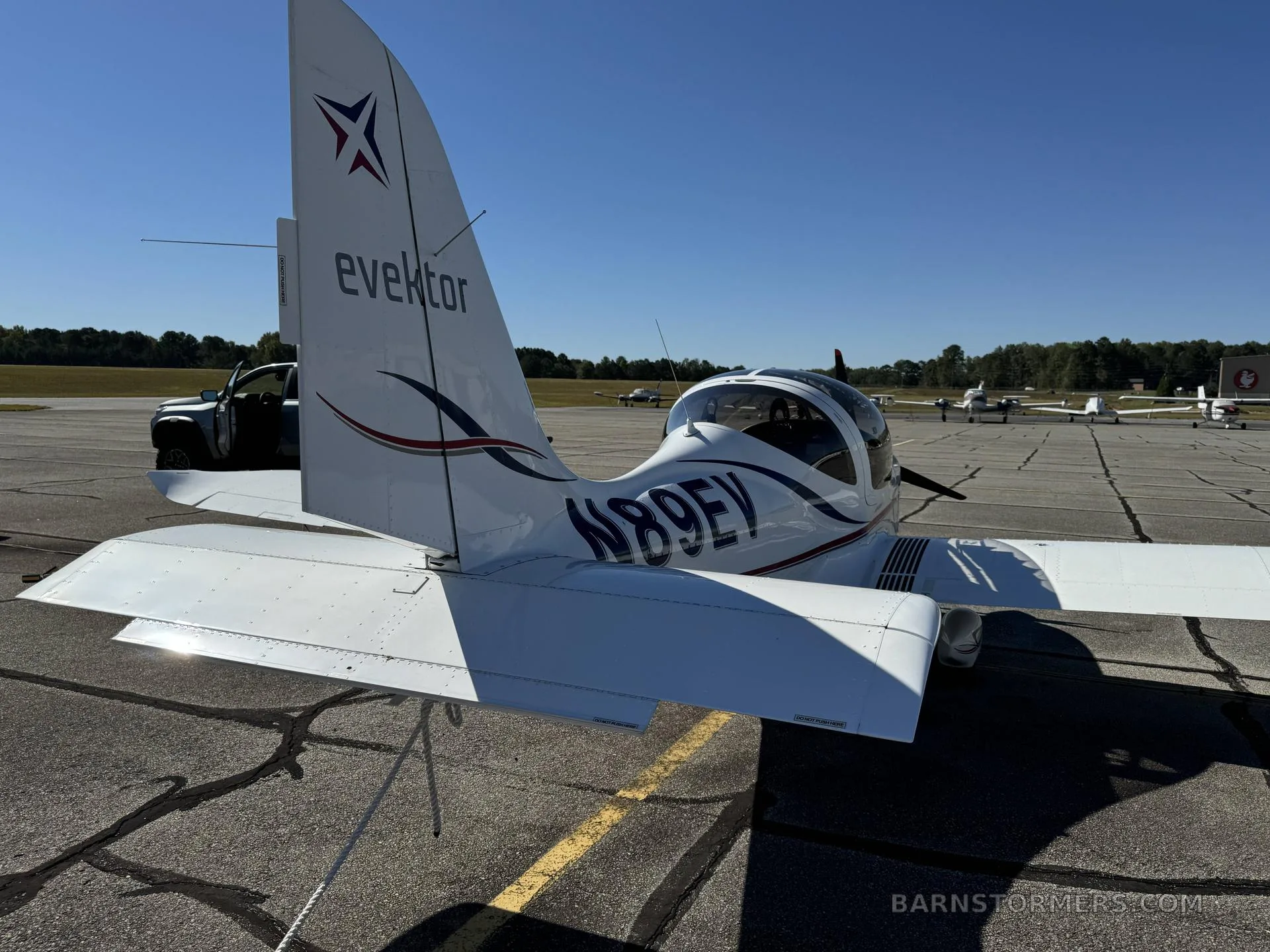 A white Evektor aircraft (N89EV) with blue and red stripes parked on an airfield, viewed from the rear-right.