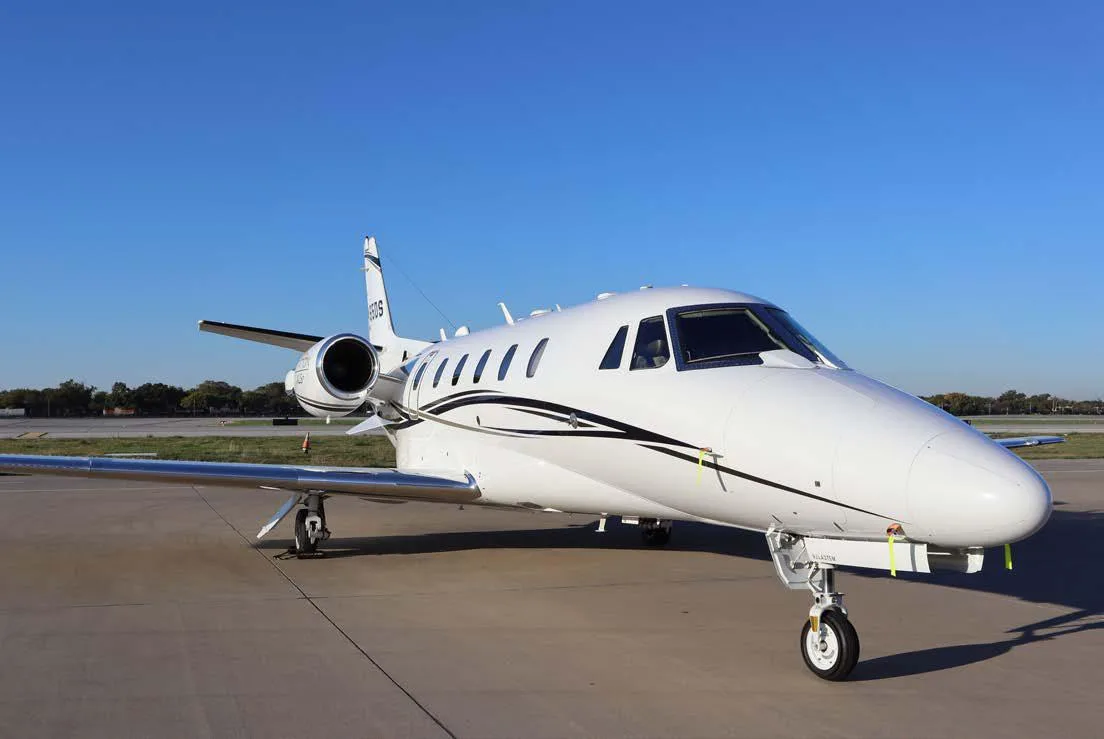 A white business jet with black and gold accent stripes is parked on an airport tarmac under a clear blue sky, viewed from the front-right.