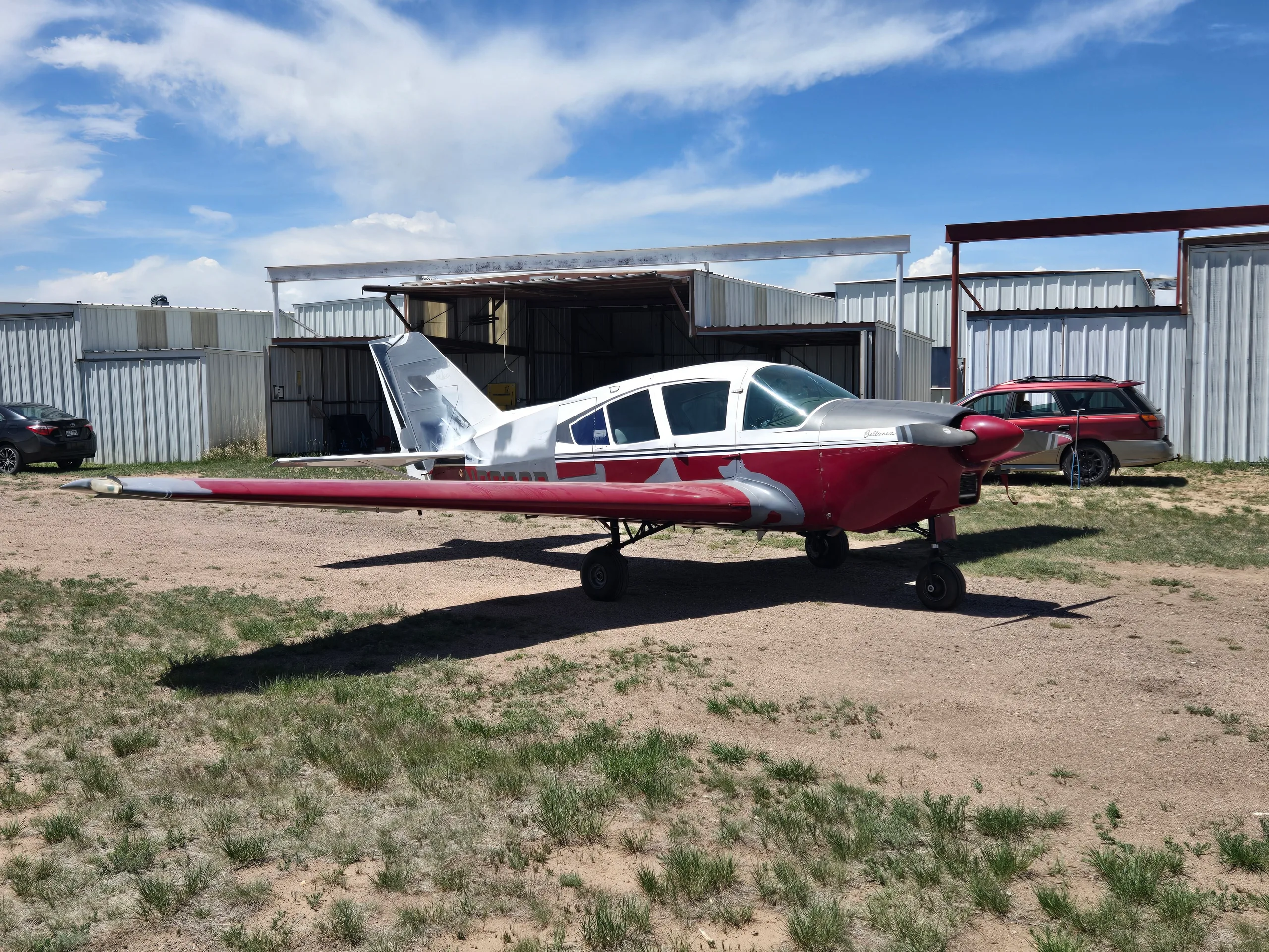 A red and silver single-engine aircraft parked on a dirt lot with multiple hangars and a red car in the background.
