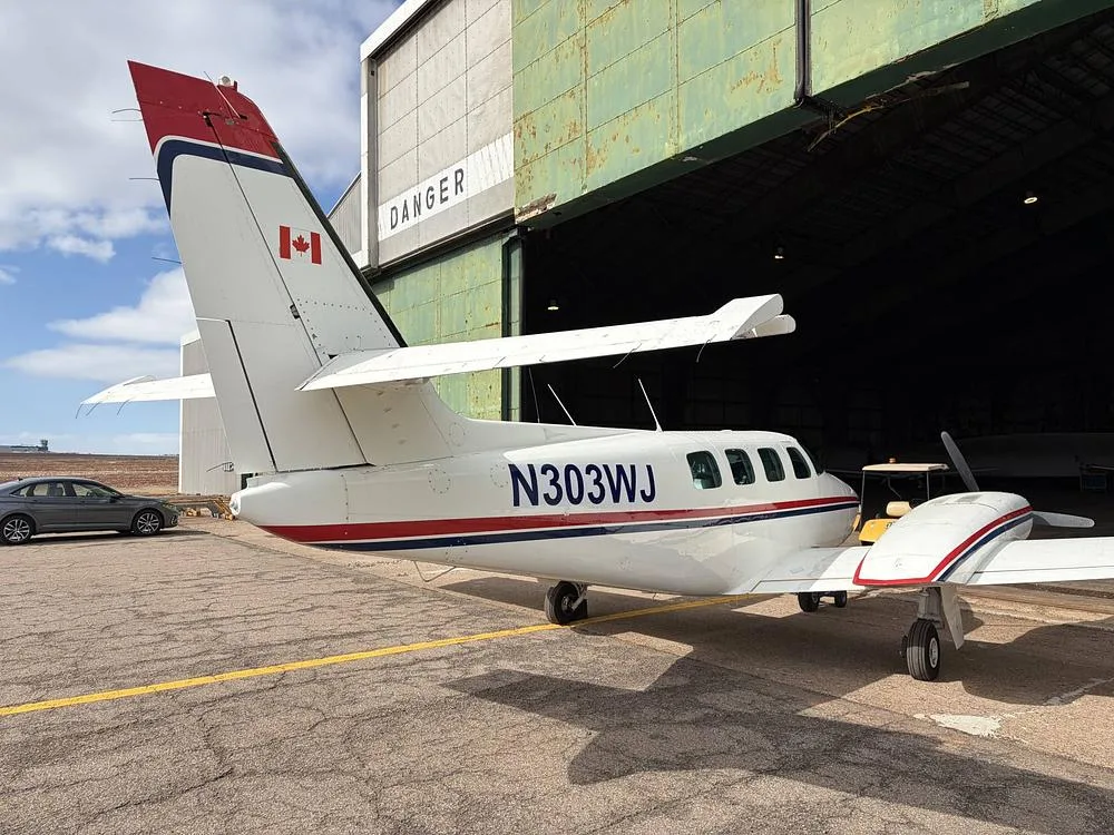 A rear-side view of the N303WJ twin-engine aircraft, showing its tail with a Canadian flag, parked near a hangar.