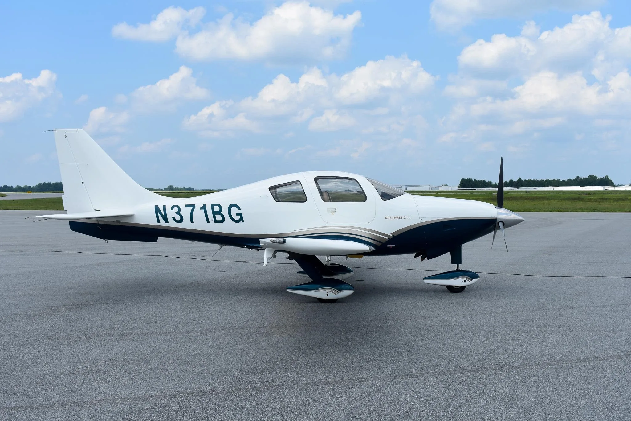 A white and blue Columbia 400 aircraft, registration N371BG, parked on an asphalt tarmac under a partly cloudy sky.