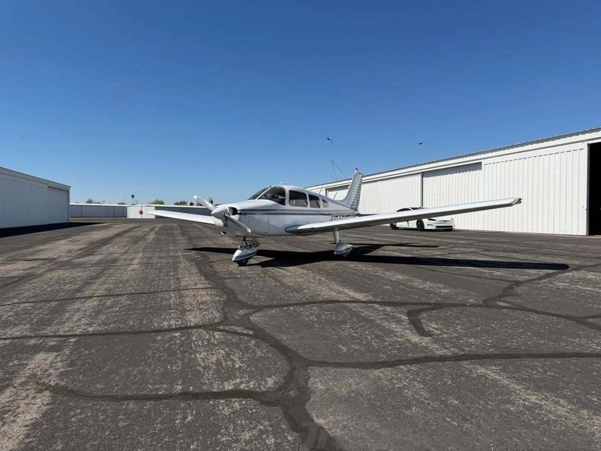A white Piper Warrior II aircraft with gray and blue stripes parked on an asphalt apron in front of hangars.
