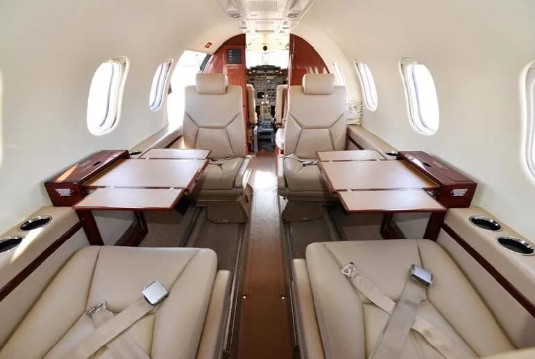 An aircraft cabin interior with light tan leather seats and fold-out tables extended, viewed looking towards the cockpit.