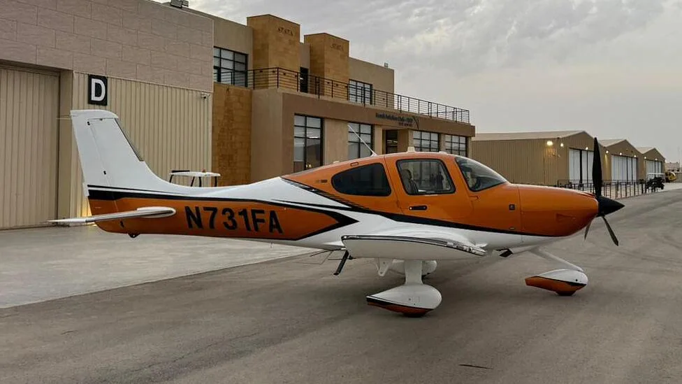 A side view of an orange and white Cirrus SR22 aircraft with registration N731FA parked on an airfield next to buildings.