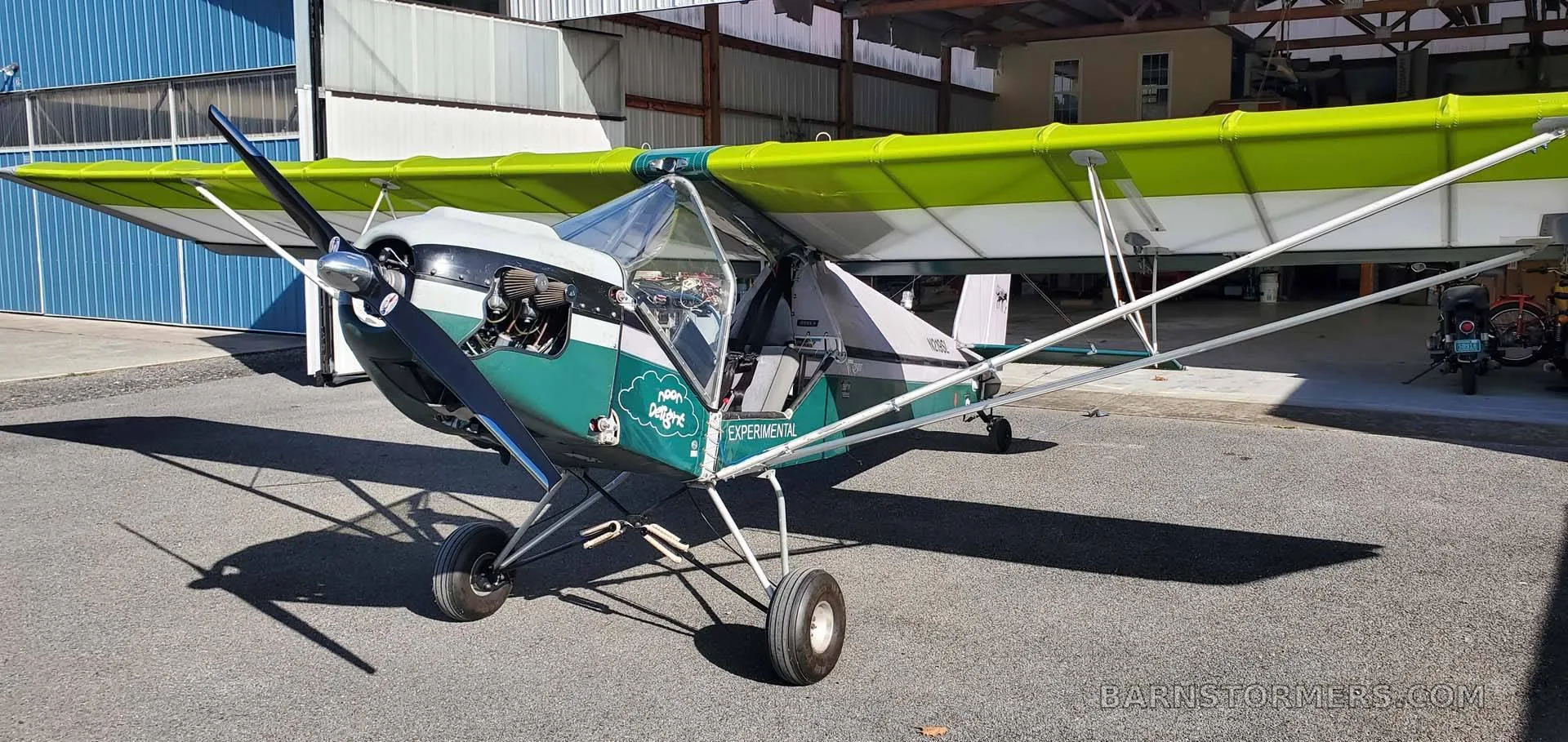 A light green and white experimental aircraft with an open cockpit, parked on asphalt in front of a hangar.