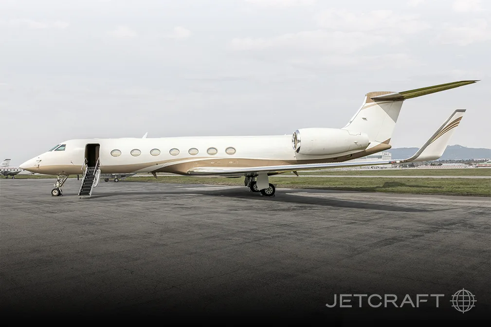 A white and gold Gulfstream G650 business jet with its door open and stairs extended, parked on an asphalt tarmac.