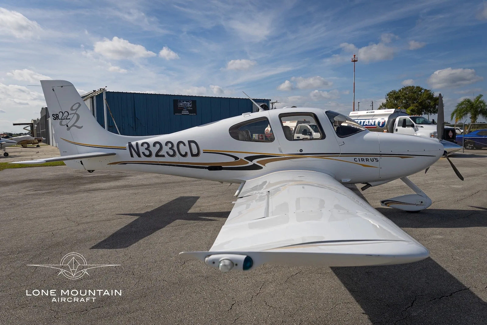 A white Cirrus SR22 aircraft with gold and black accents, registration N323CD, parked on an airfield next to a blue hangar.