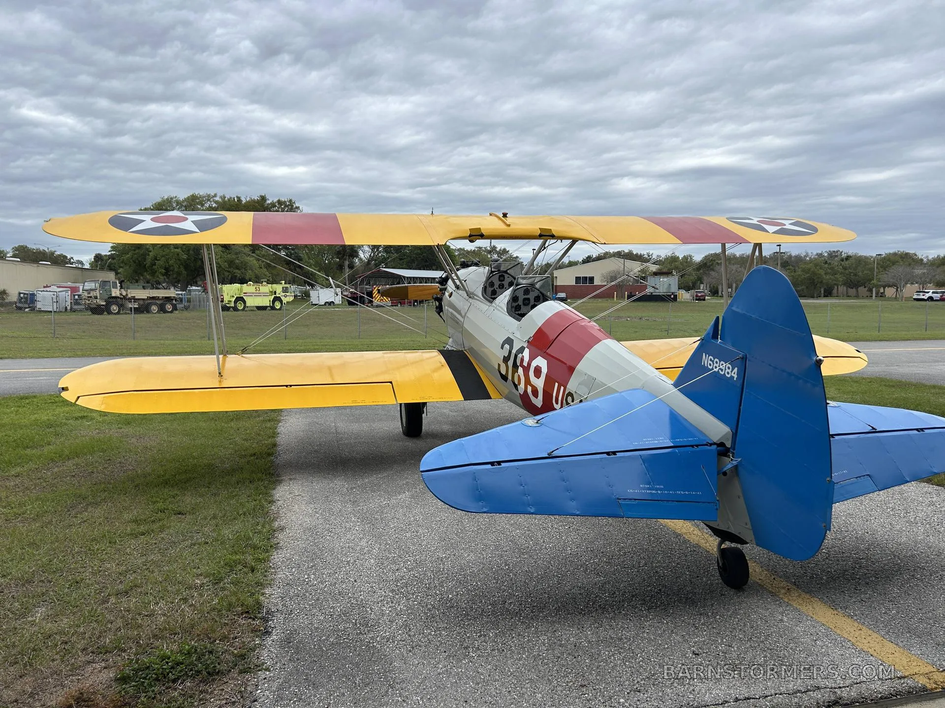 A yellow and white biplane with 'US NAVY 369' markings and a blue tail, seen from the rear on an asphalt ramp with trees in the background.