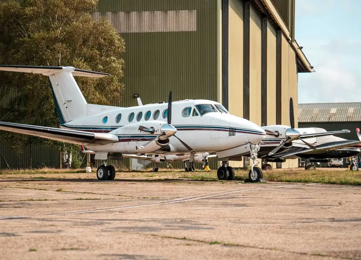 A white twin-turboprop aircraft with dark stripes parked on an asphalt apron next to hangars.