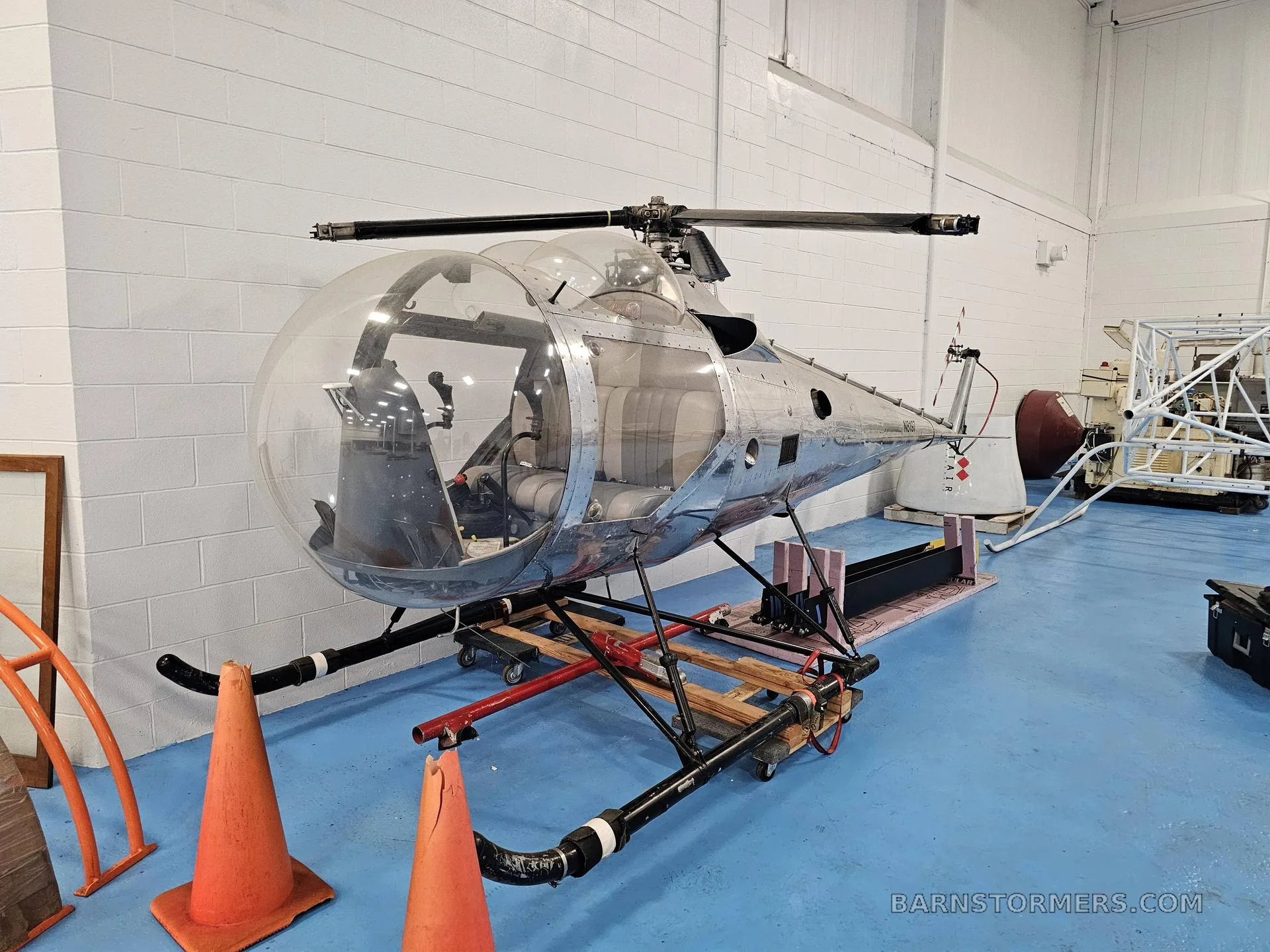 A shiny silver helicopter with a clear bubble canopy is shown from the front-left, resting on a wooden stand in a hangar.