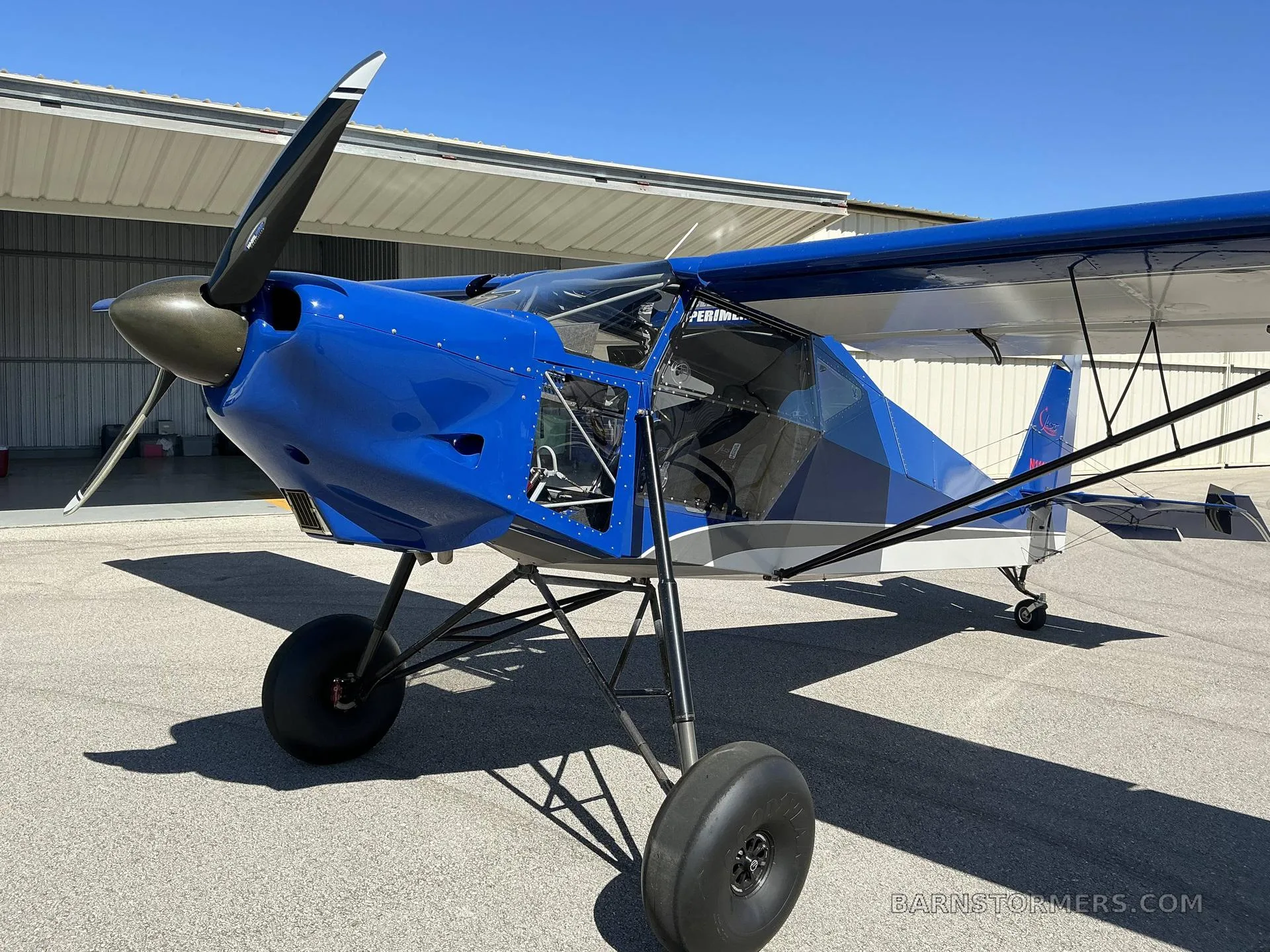 A front-quarter view of a blue and white SuperSTOL aircraft with its propeller visible, parked on an asphalt apron.
