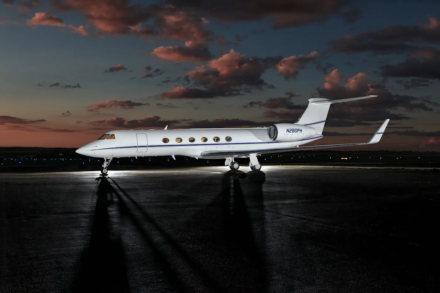 A white business jet with a blue stripe, registration N280PH, parked on a tarmac at sunset with dramatic clouds.