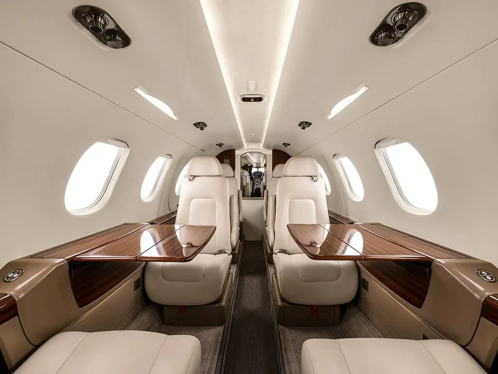The interior cabin of an Embraer Phenom 300, showing light cream leather seats with dark wood tables extended.