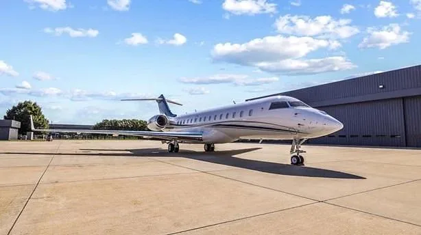 A white business jet with blue tail accents is parked on a tarmac in front of a hangar under a partly cloudy sky.