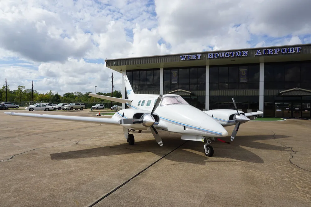 A white twin-engine propeller aircraft with blue stripes, registration N818KT, parked on the tarmac in front of the West Houston Airport building.