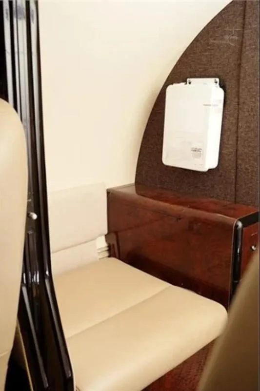Close-up view of a beige leather seat and a dark wood panel with a white electronic device mounted on the wall inside the aircraft cabin.