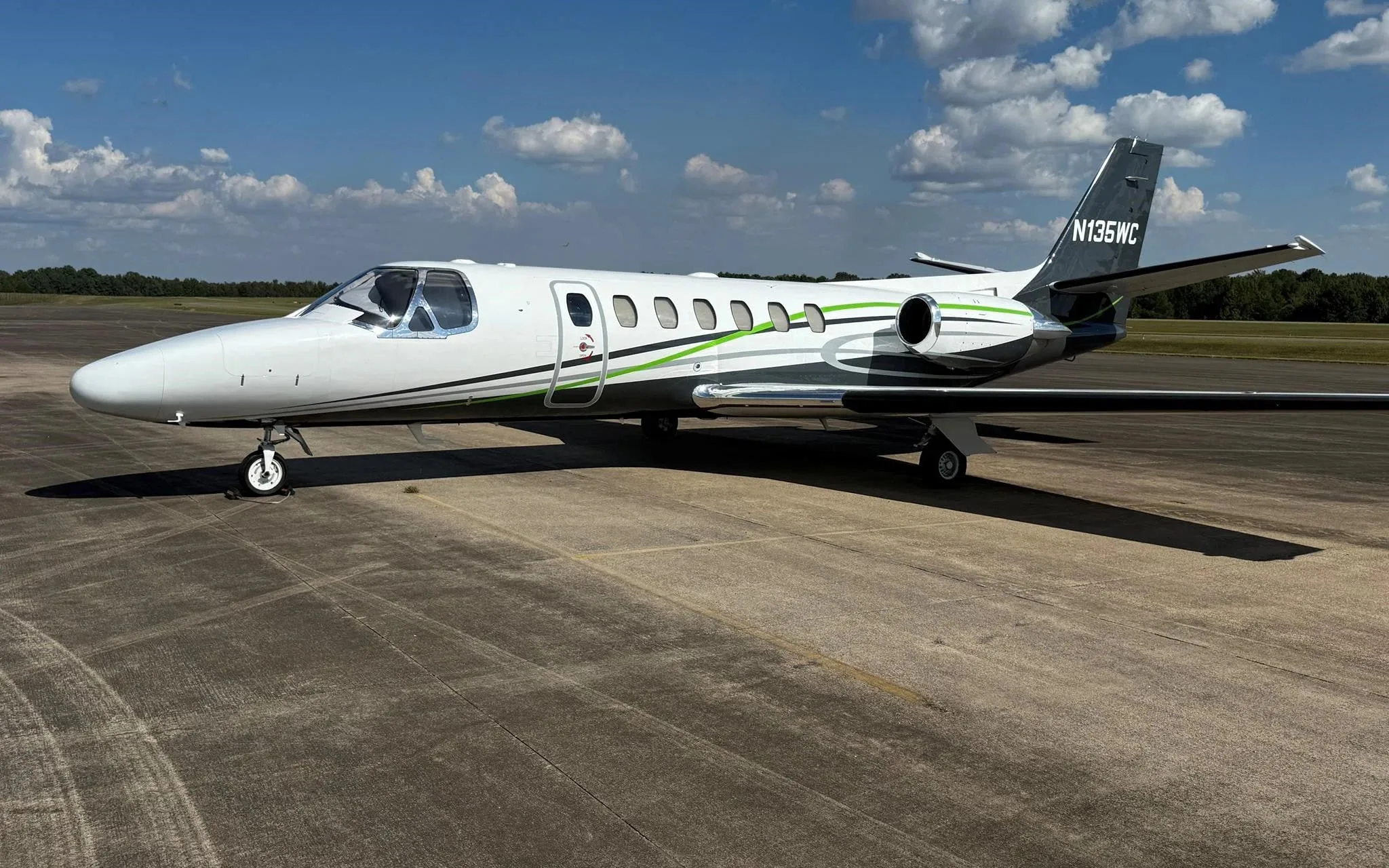 A white and grey business jet with green accents, registration N135WC, parked on an asphalt tarmac under a partly cloudy sky.