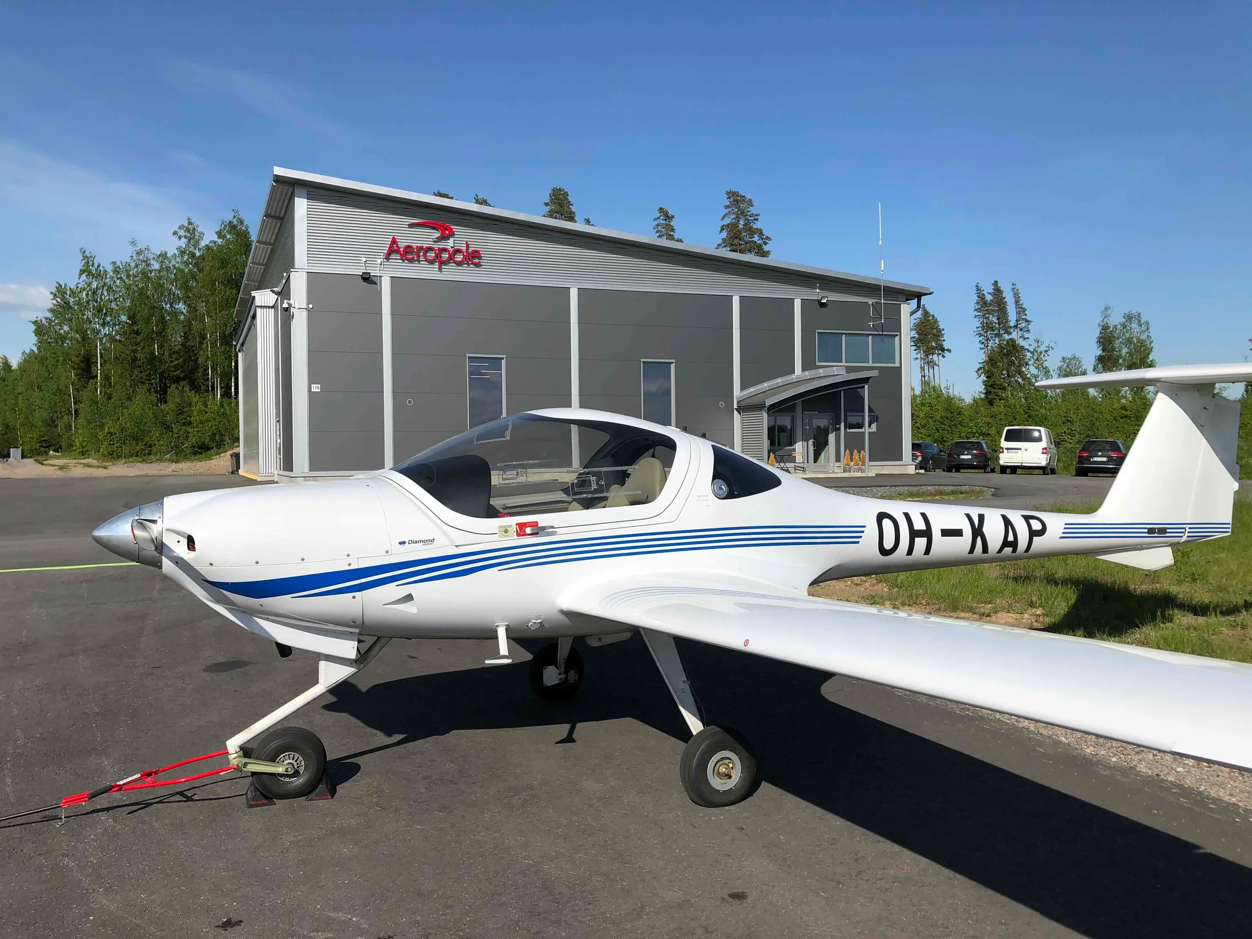 A white Diamond DA20 aircraft with blue stripes is parked on an airfield tarmac in front of a grey hangar under a clear sky.