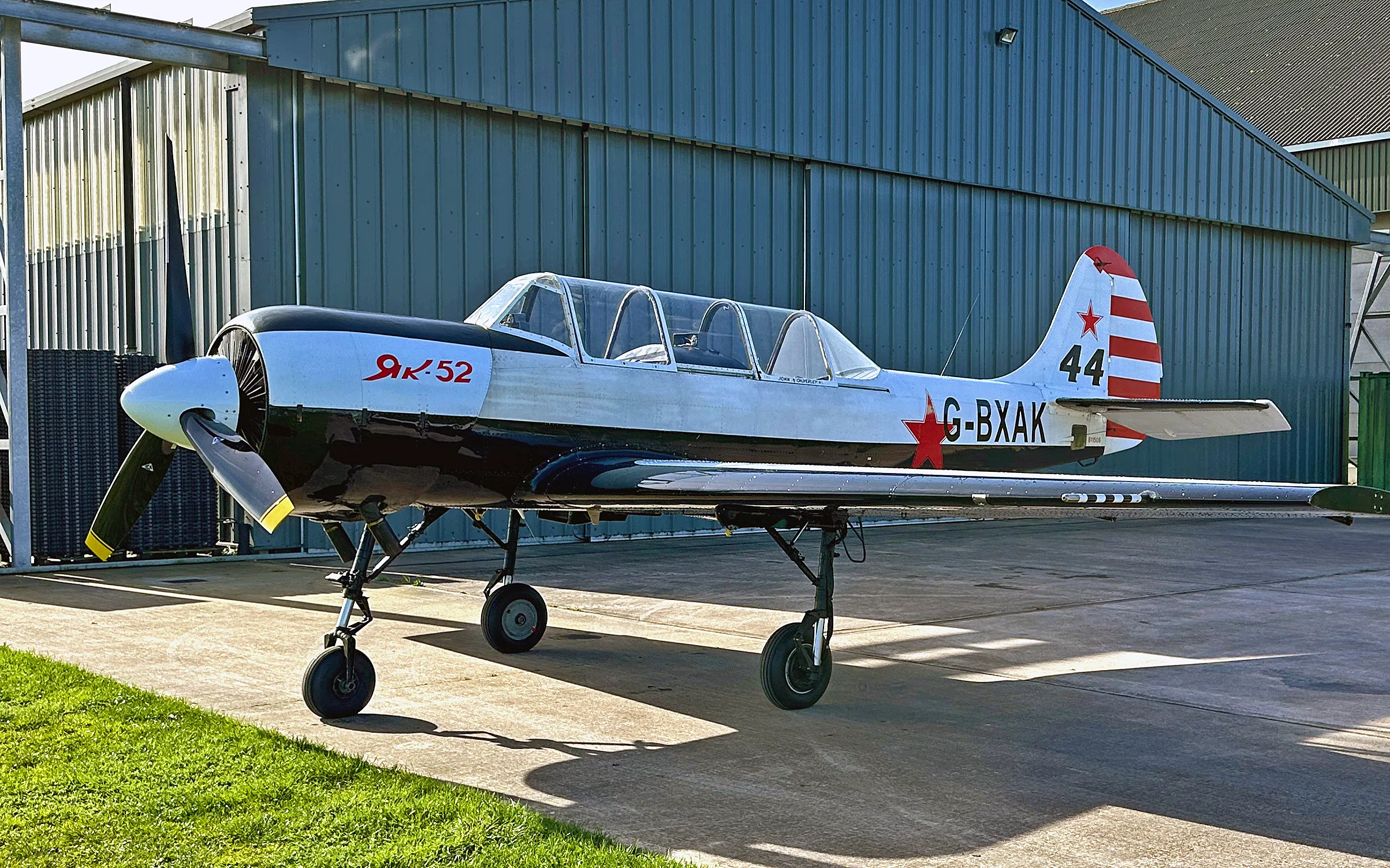 A white and black Yak-52 aircraft with red stars and tail stripes, parked on a concrete pad in front of a blue hangar.
