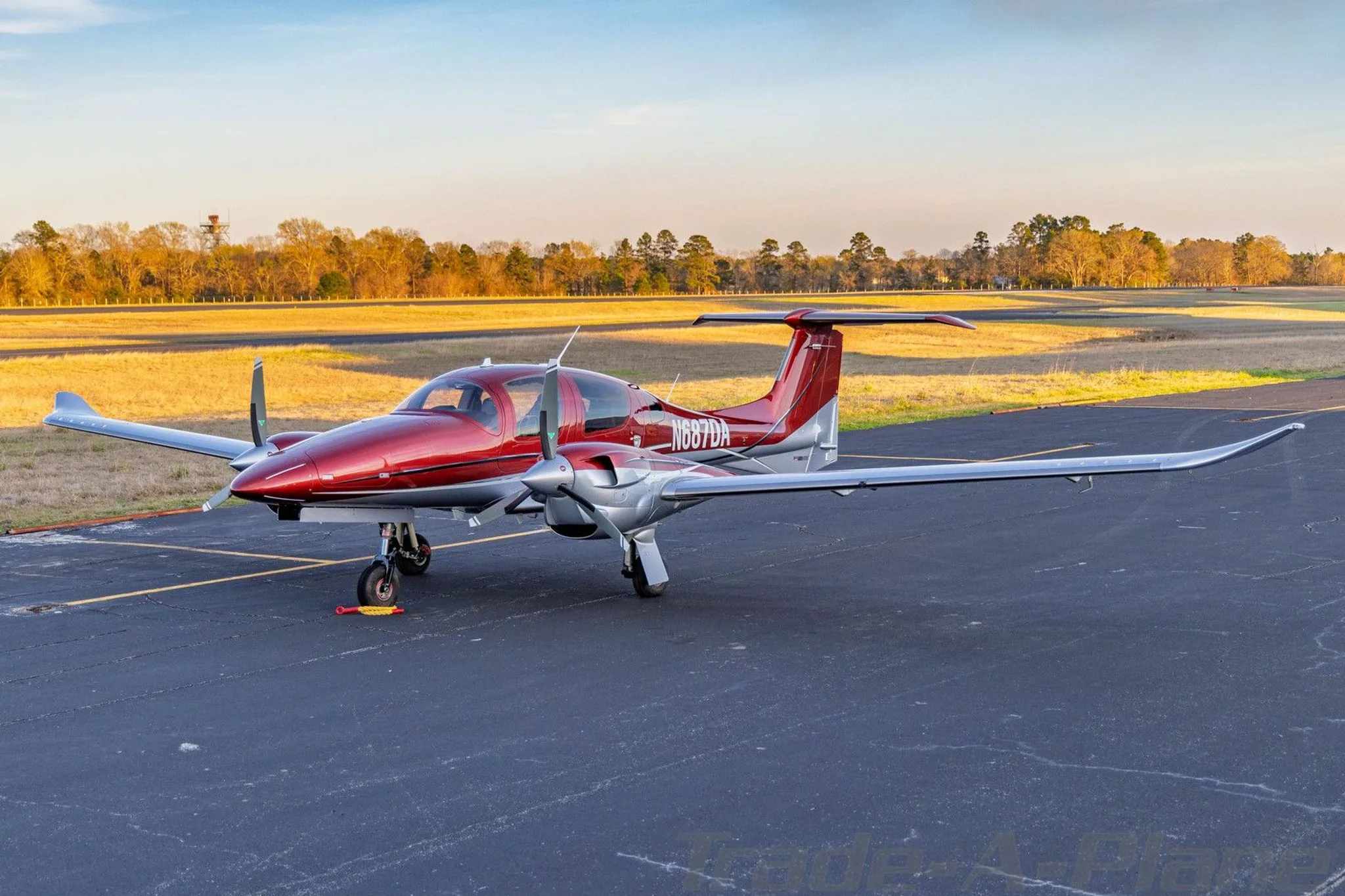 A red and silver Diamond DA62 aircraft parked on an airfield at sunset.