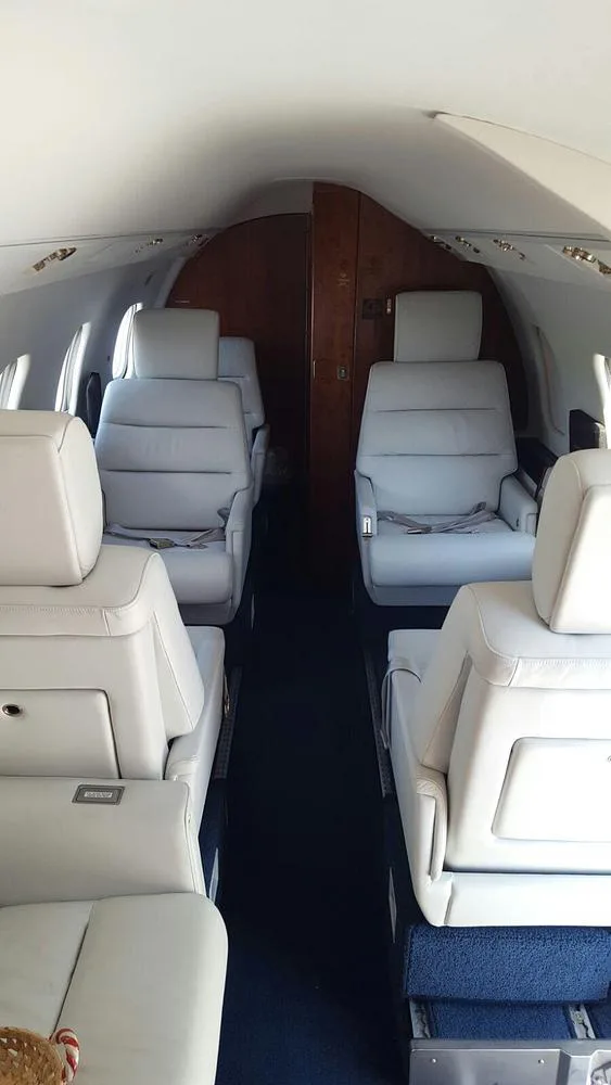 The interior cabin of a private jet with light grey leather seats and a dark blue carpet, looking towards the rear.