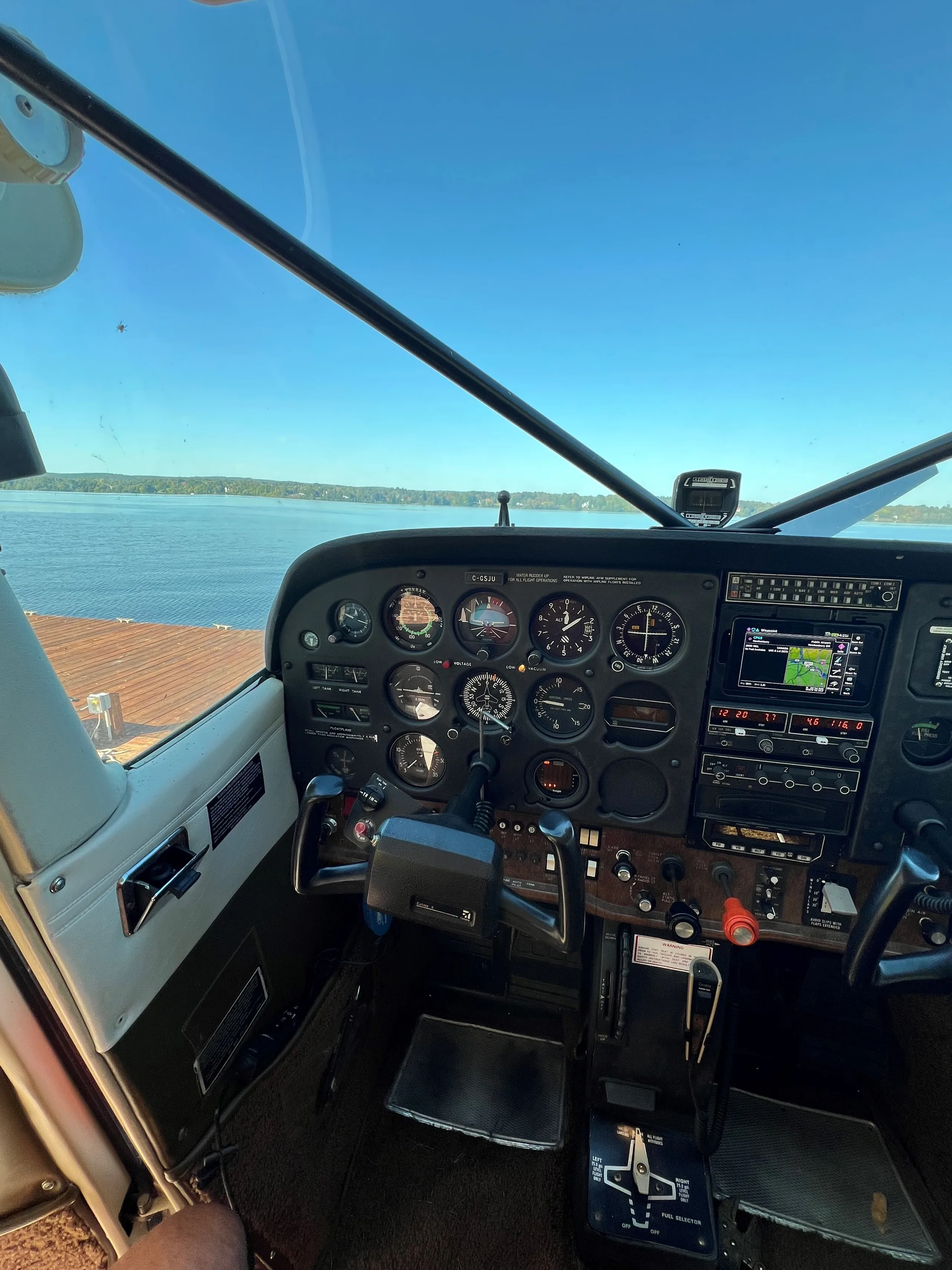 The cockpit of a floatplane shows the instrument panel and controls, with a view of a lake outside.