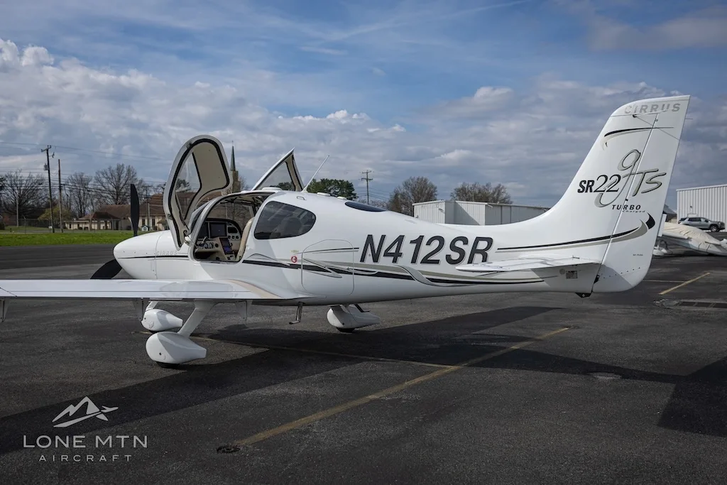 A white Cirrus SR22T aircraft with open doors is parked on an asphalt runway under a partly cloudy sky.