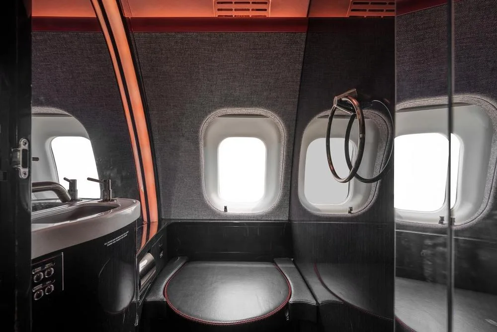 A modern aircraft lavatory features a sink, toilet, and two windows, with dark grey walls and red accent lighting.