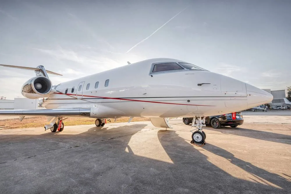 A white business jet with red and black accent stripes parked on an asphalt tarmac under a bright sky, viewed from the front-right.