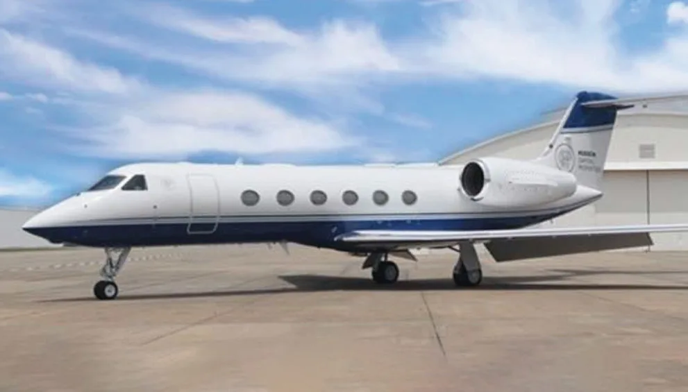 A white and blue Gulfstream business jet is parked on an airfield in front of a hangar under a partly cloudy sky.