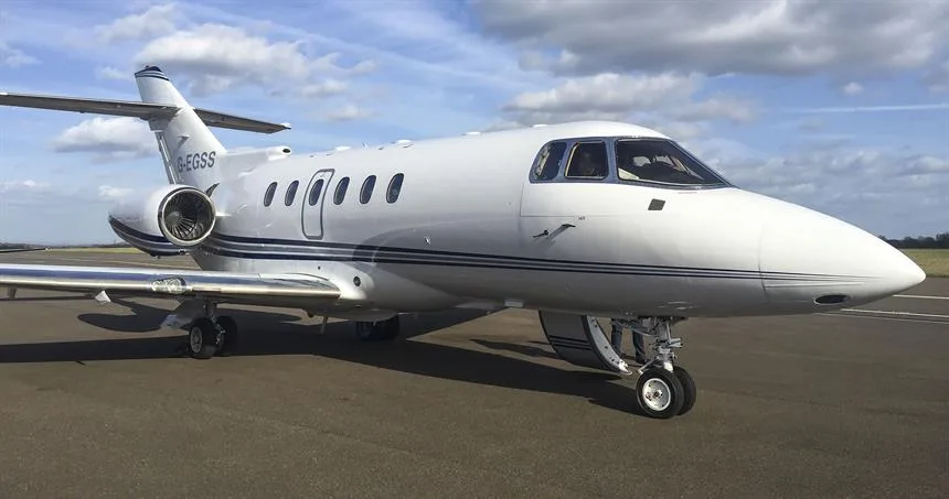 A white business jet with blue stripes, registration G-EGSS, parked on an airfield under a partly cloudy sky.