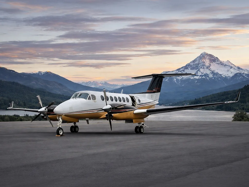 A white and gold twin-turboprop aircraft, likely a King Air, parked on a tarmac with scenic mountains and a sunset sky in the background.