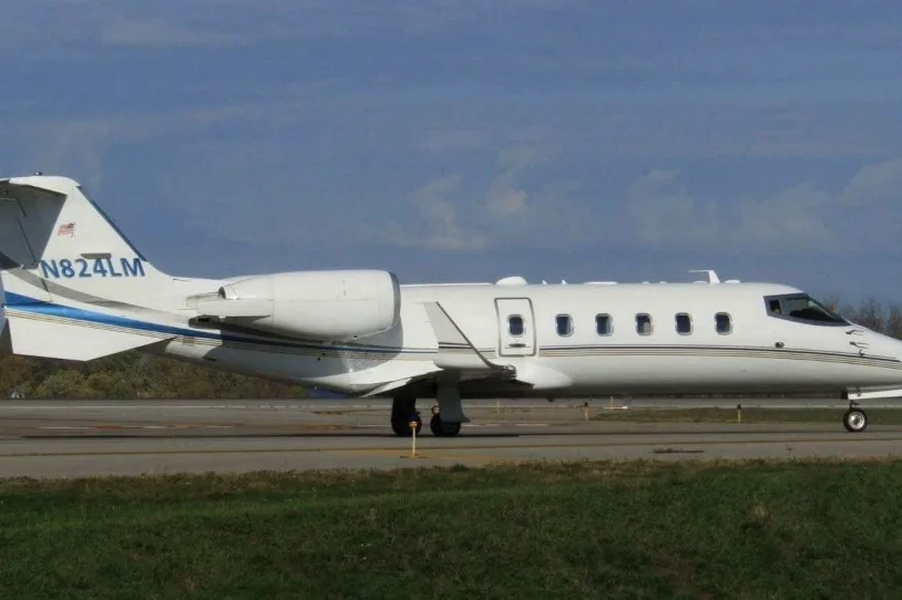 A white business jet with blue and gold stripes, registration N824LM, on a tarmac under a partly cloudy sky.