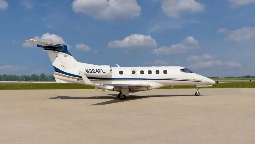 A side view of a white Embraer Phenom 300 private jet with blue and gold stripes, parked on an airfield.