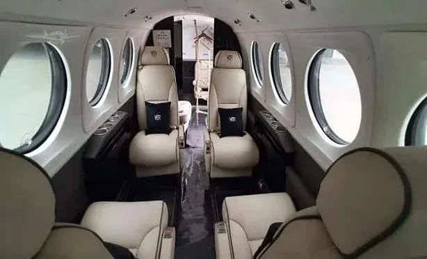 The aircraft cabin shows rows of light-colored leather passenger seats and large oval windows.