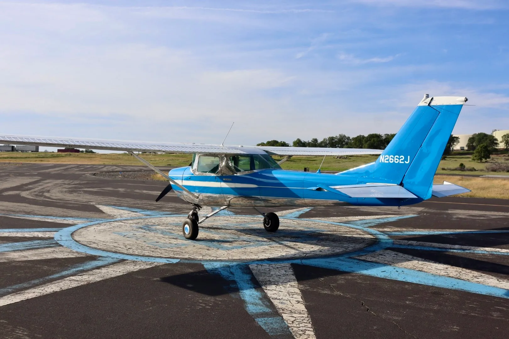 A blue and silver Cessna 150 aircraft with registration N2662J parked on a compass rose at an airfield.
