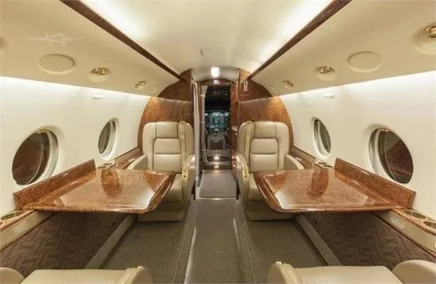 The luxurious interior of a private jet cabin featuring two beige leather seats with wood-grain tables and a central aisle.
