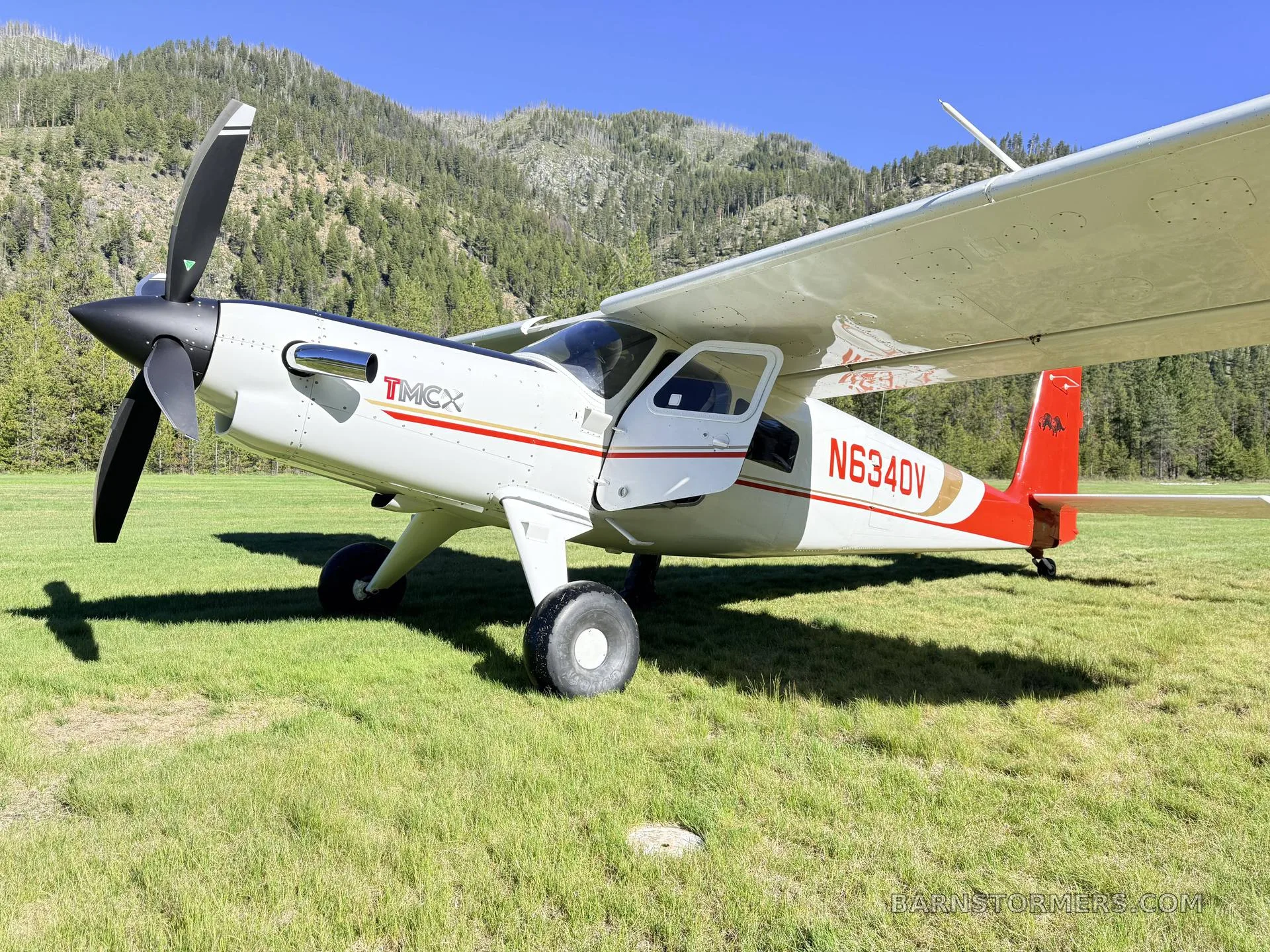 A white and red T-MCX aircraft (N634DV) with a black propeller is parked on a grassy field with mountains in the background.